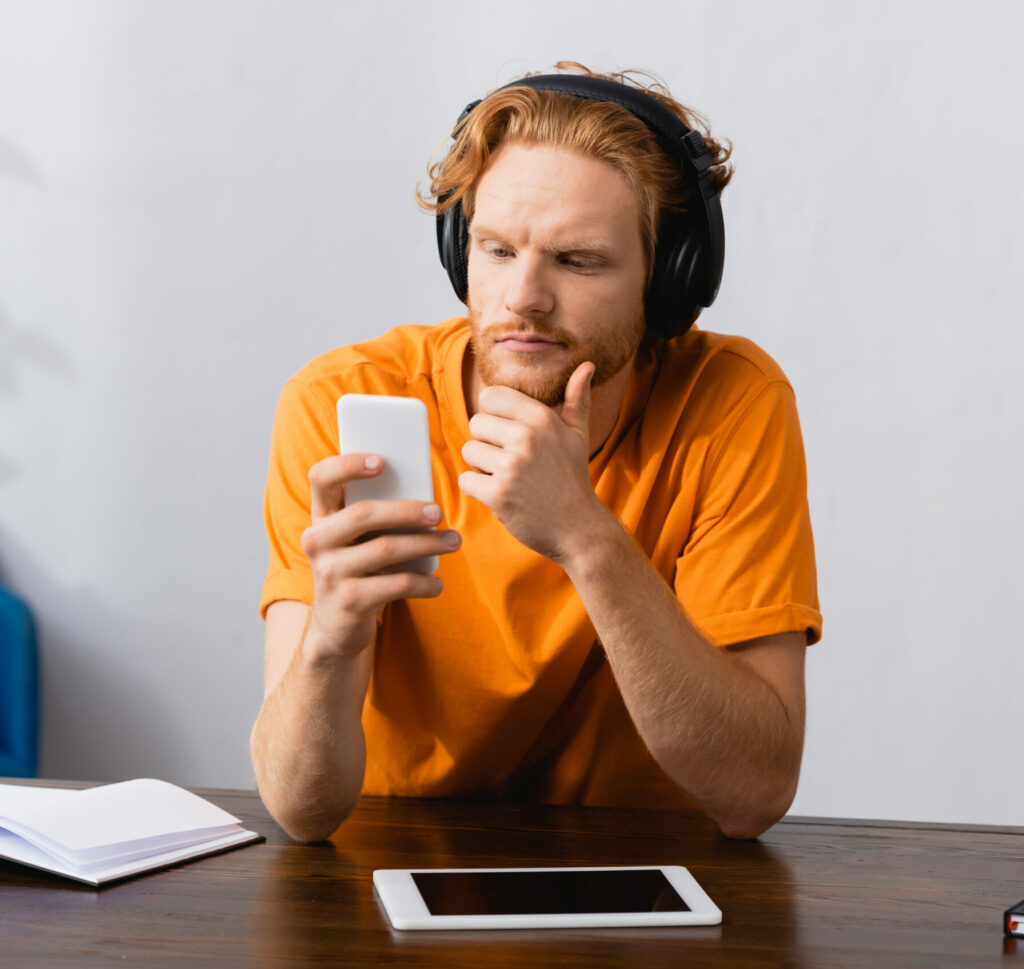 Am Tisch sitzender Mann mit Kopfhörern und Smartphone in der Hand beim Podcast-Hören