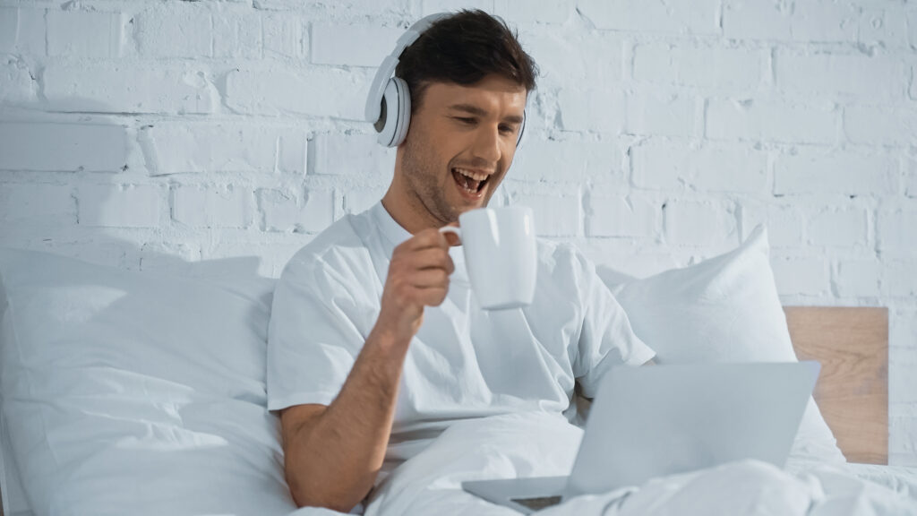 Ein erfreuter Mann mit Kopfhörern sitzt im Bett und trinkt Kaffee.