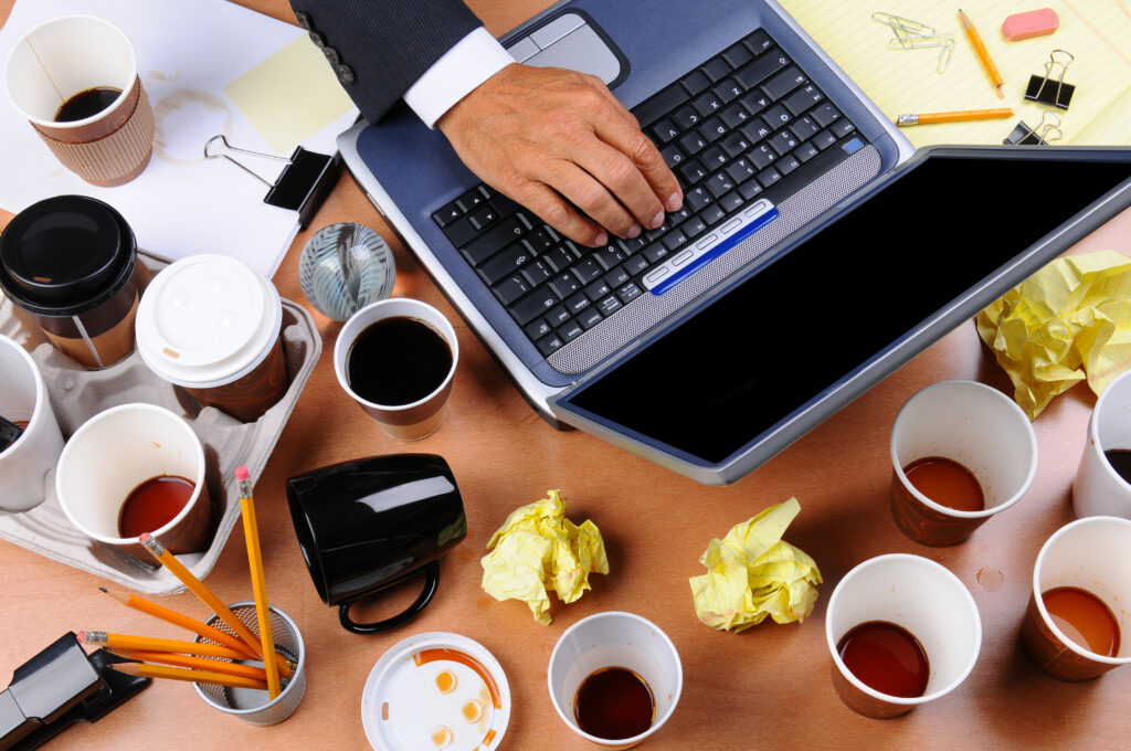 Hand von Geschäftsmann auf Laptop, umgeben von lauter leeren Kaffeebechern und zerknülltem Papier