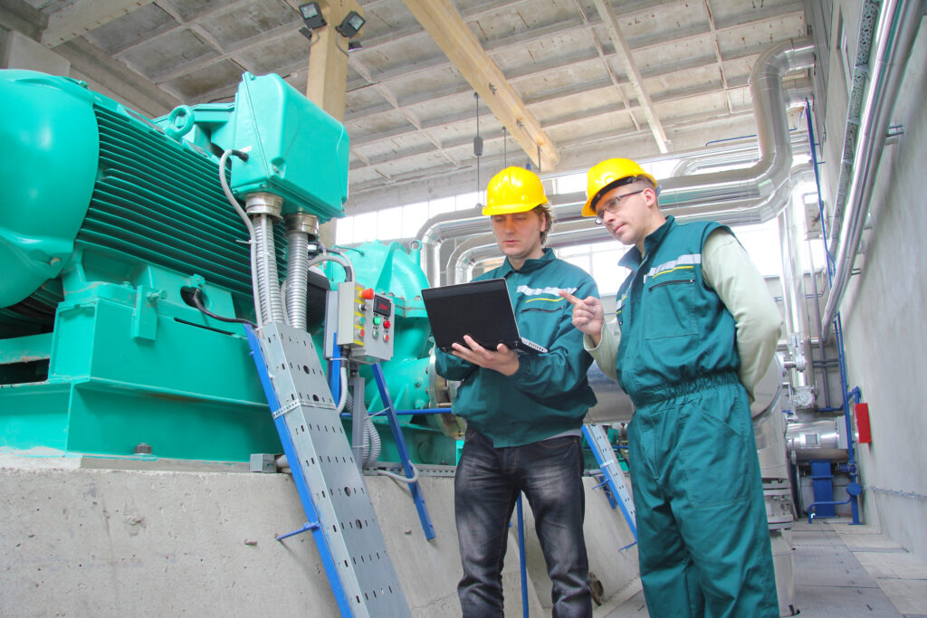 2 Ingenieure mit Schutzhelmen stehen neben einer großen Maschine in einer Fabrikhalle.